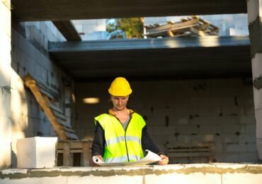 A construction engineer and designer in a yellow hardhat studies a drawing of a building